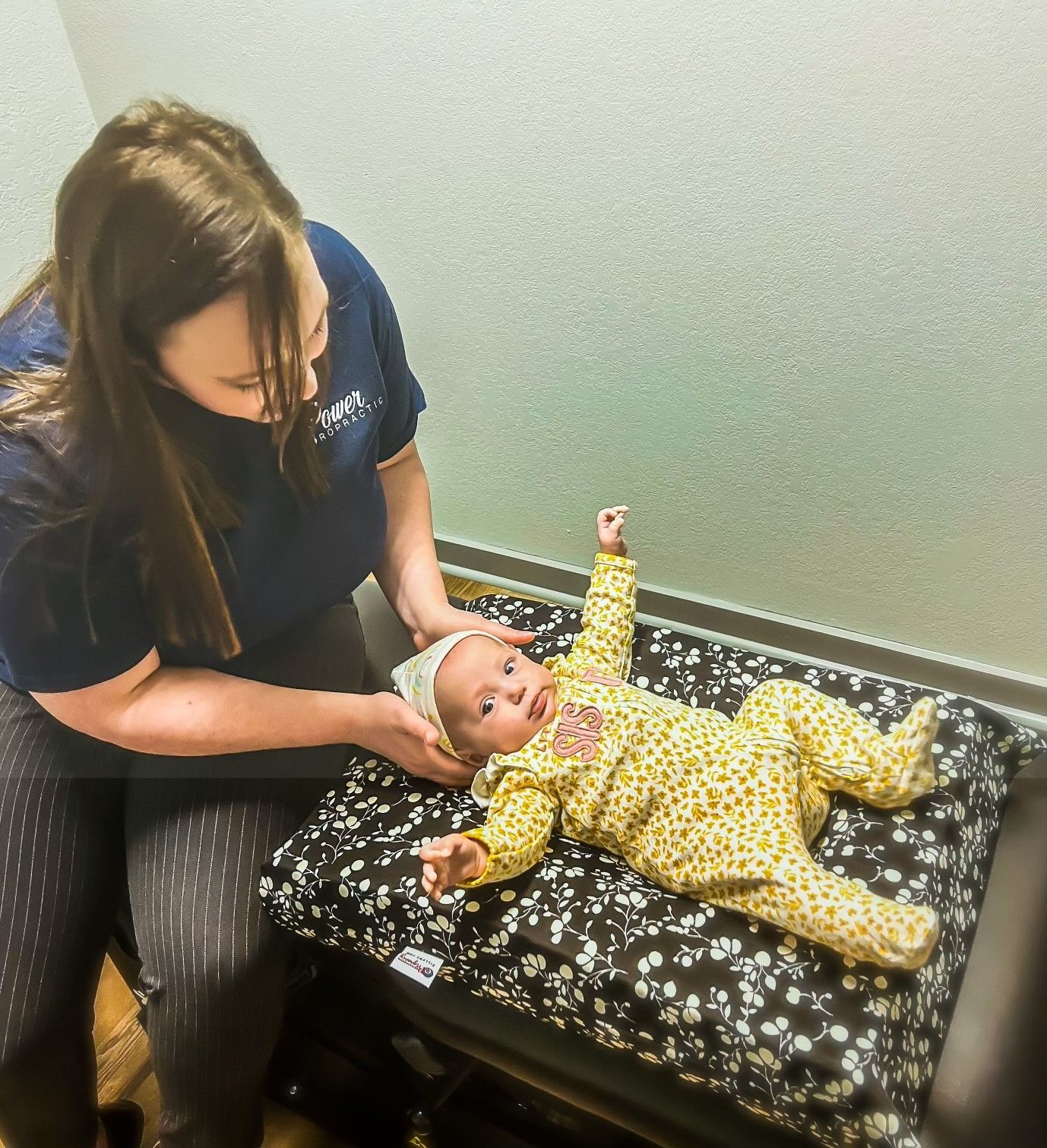 Caregiver adjusts a baby lying on a black mat, wearing a yellow patterned onesie.