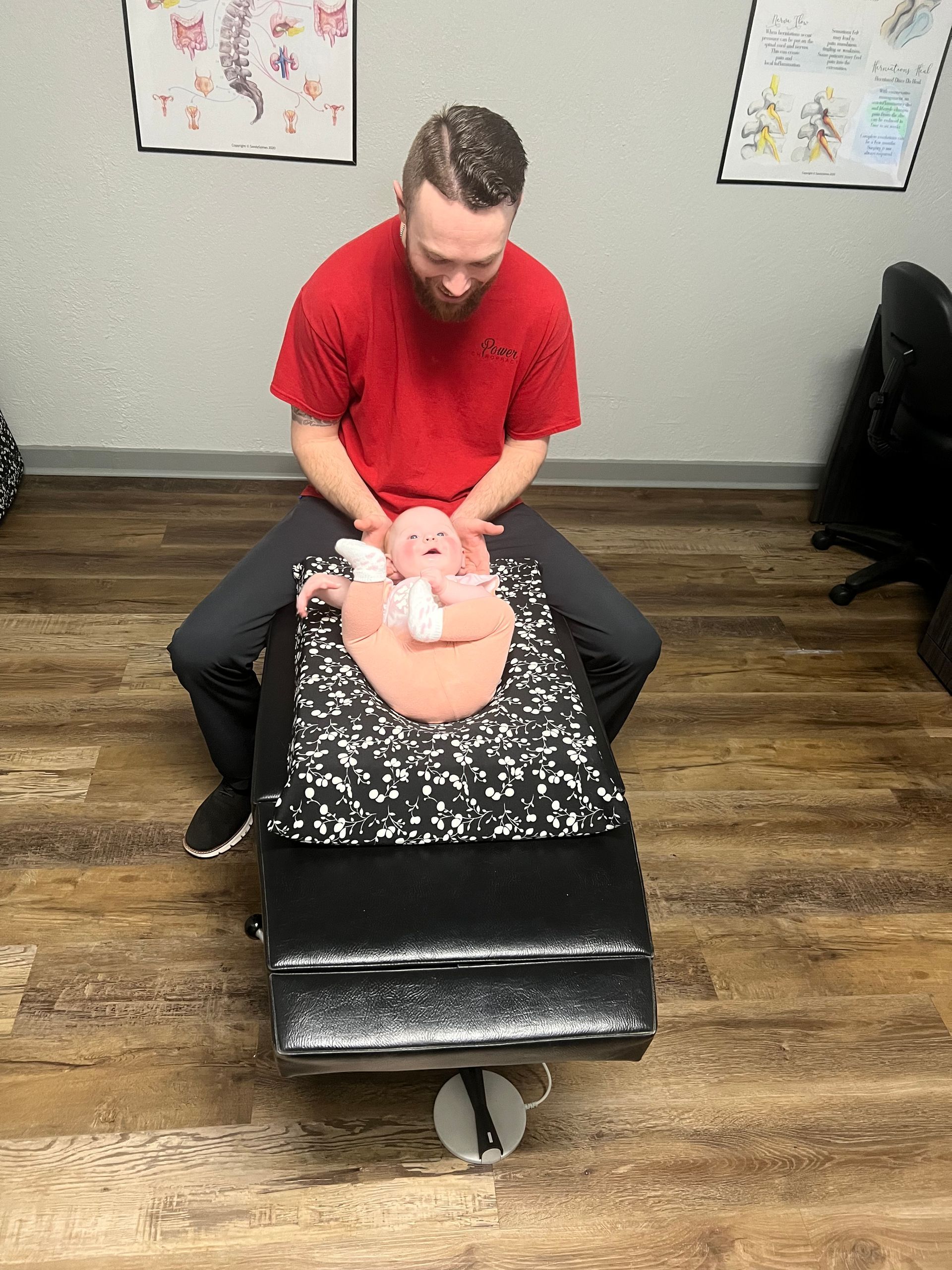 Dentist treating a patient reclined on a black chair in a clinic, with a red shirt and wood floor.