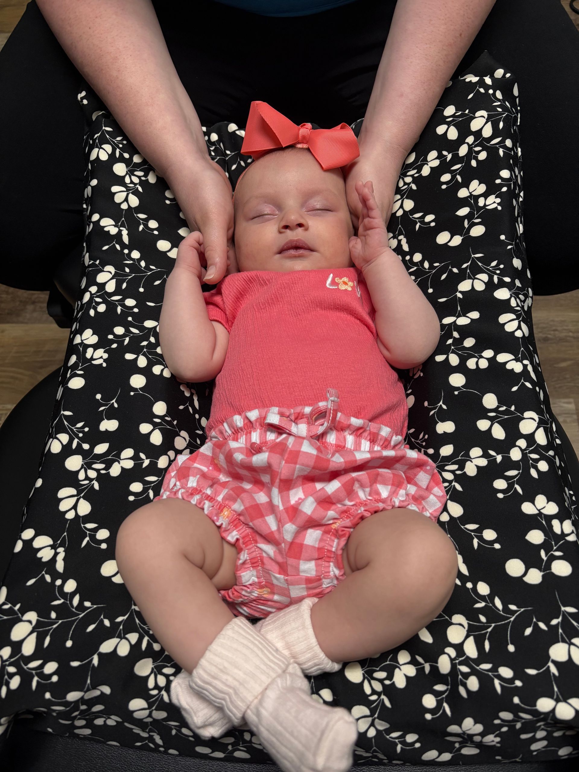 Sleeping baby in pink outfit with bow, lying on a black floral cushion and held by adult hands