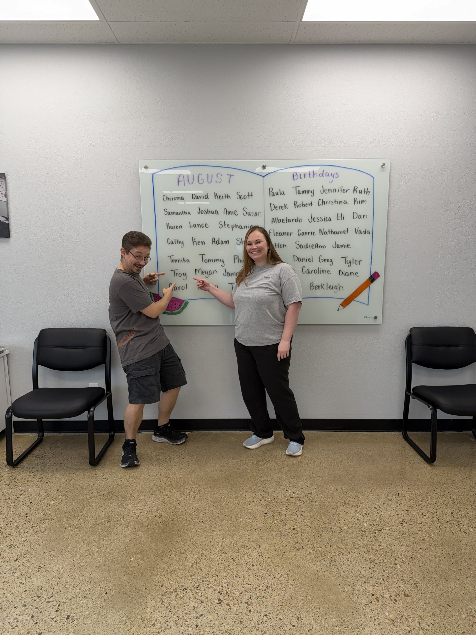 Two people pose in front of a whiteboard in a classroom, with chairs along the wall.