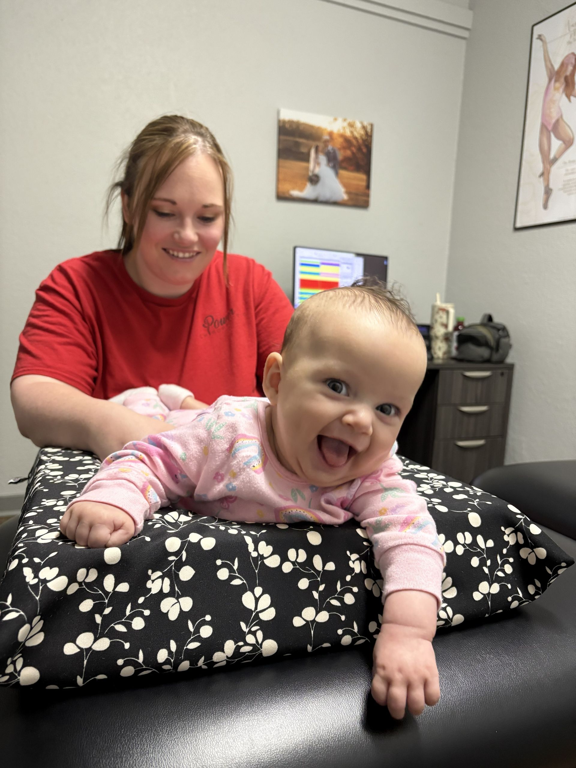 Smiling baby in pink pajamas on a black mat, with a woman in red smiling behind in a treatment room