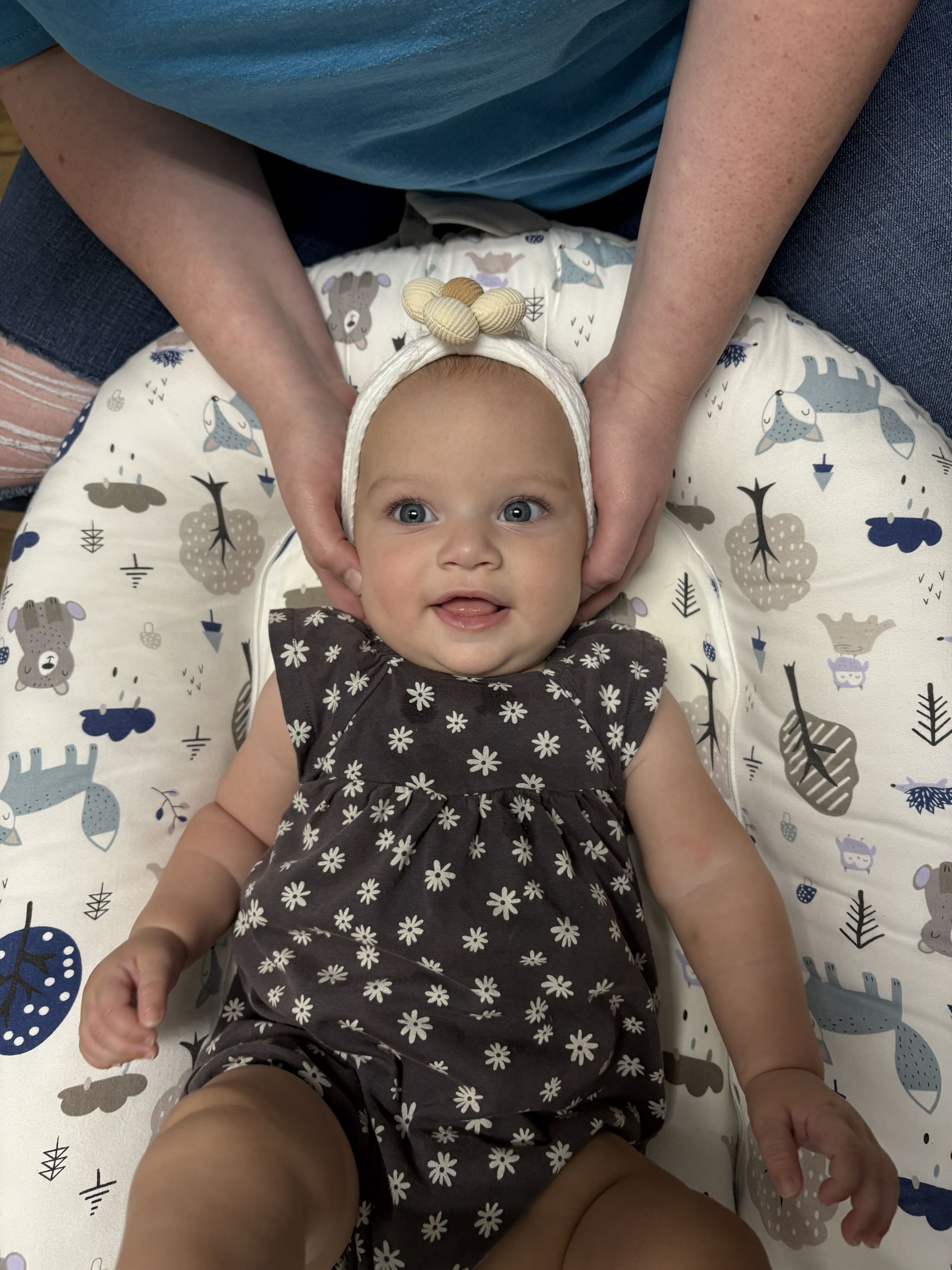 Baby lying on a patterned pillow, held gently by an adult, wearing a dark floral onesie and a headband with a bow