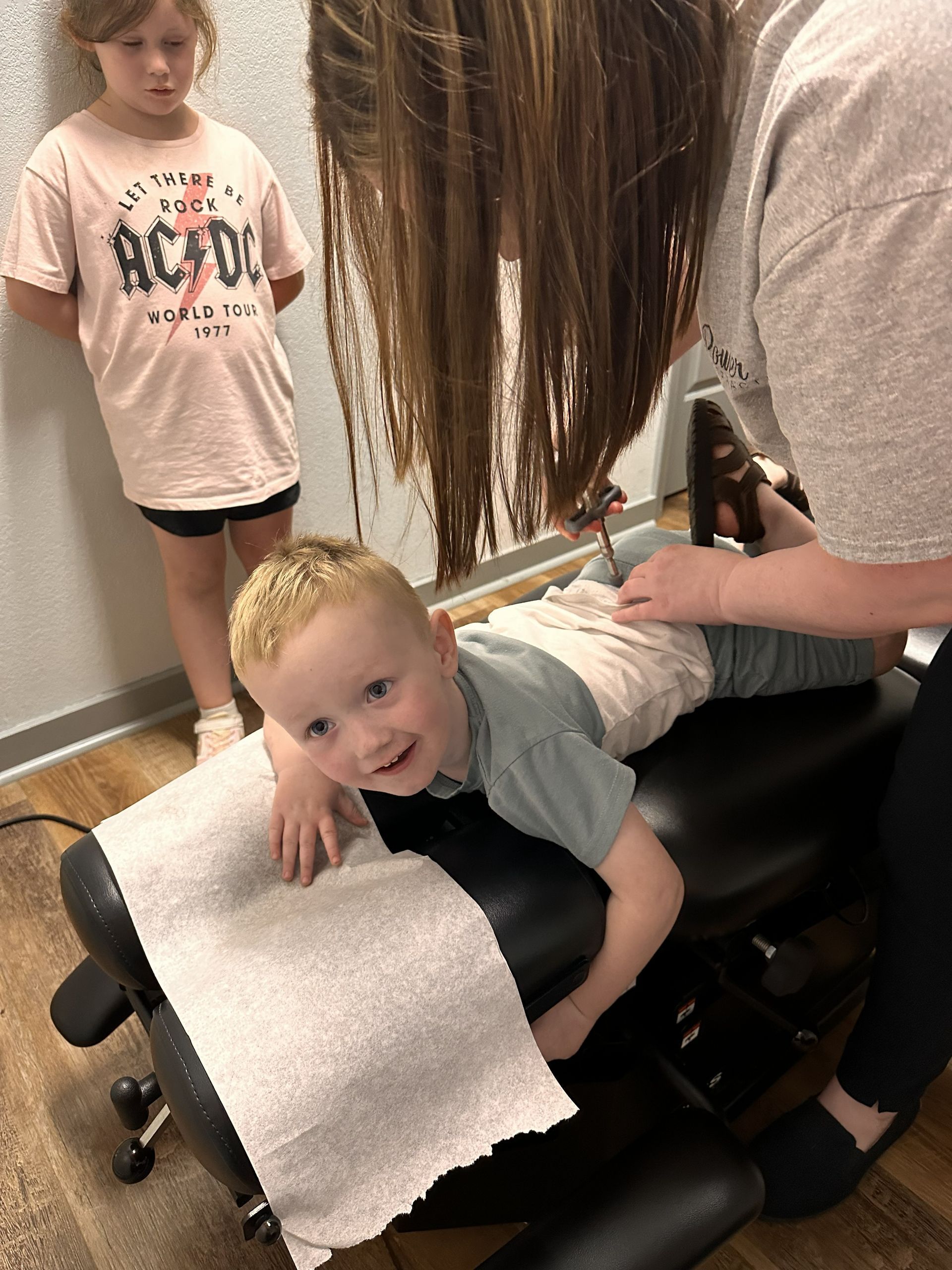 Child getting a chiropractic adjustment while another child stands nearby in a clinic room