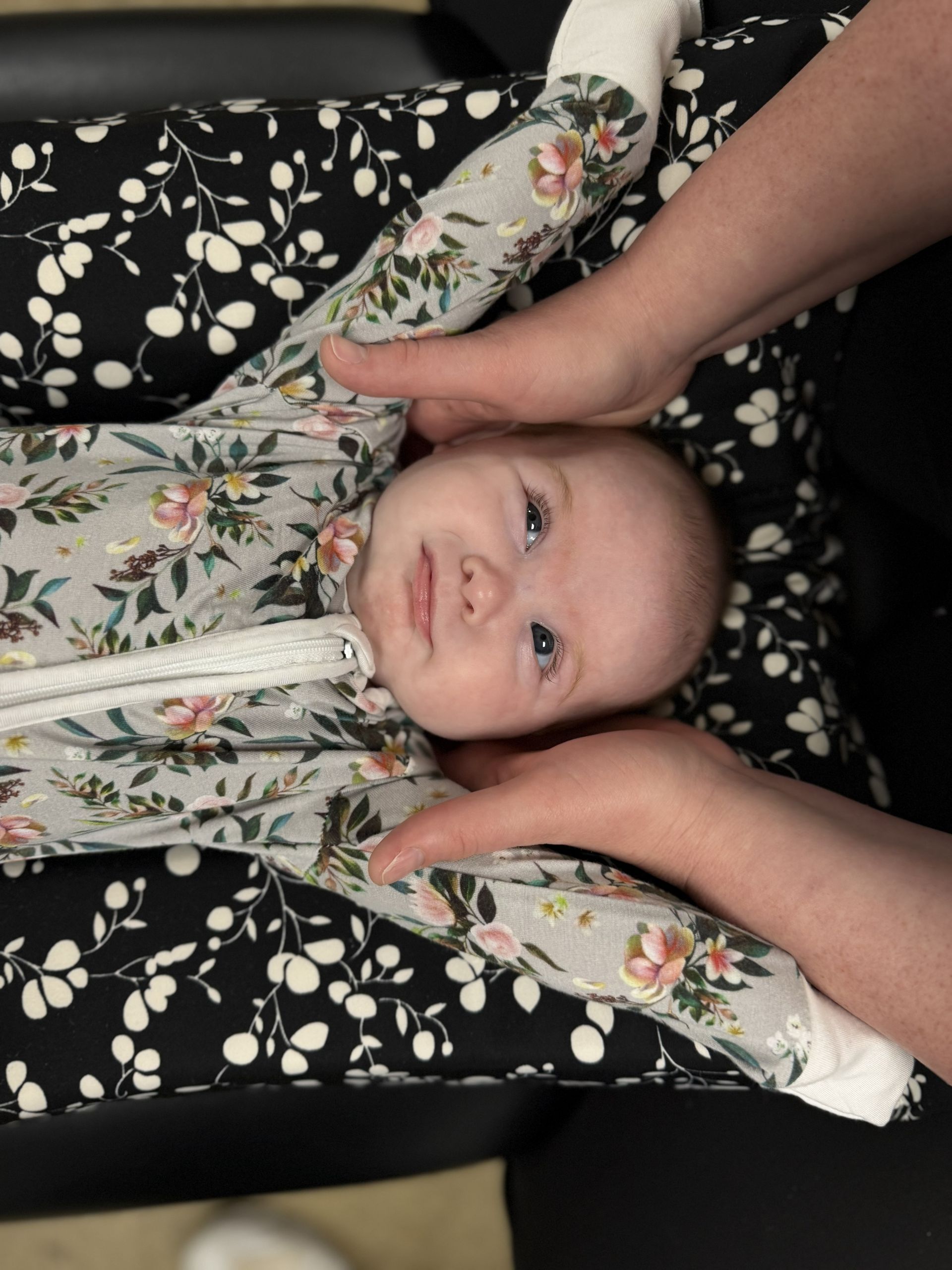Baby smiling while lying in a floral car seat liner, with hands gently holding their head