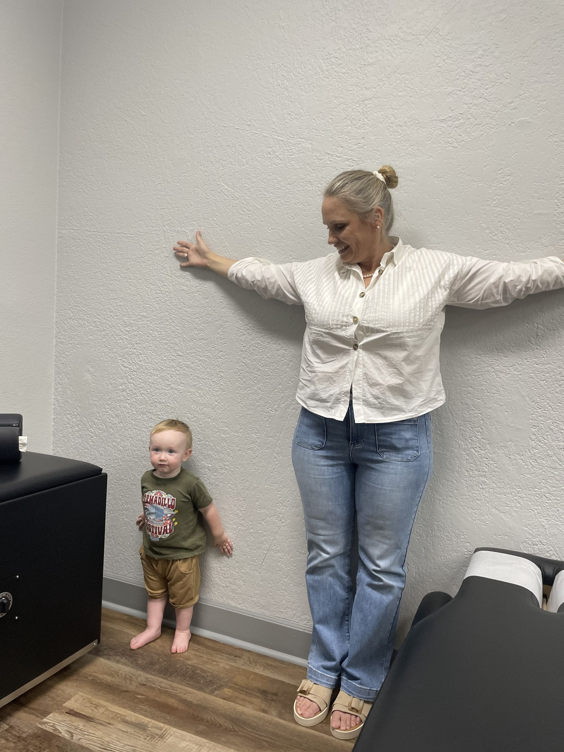 Woman and toddler standing against a wall in an office, arms outstretched; both facing the camera.