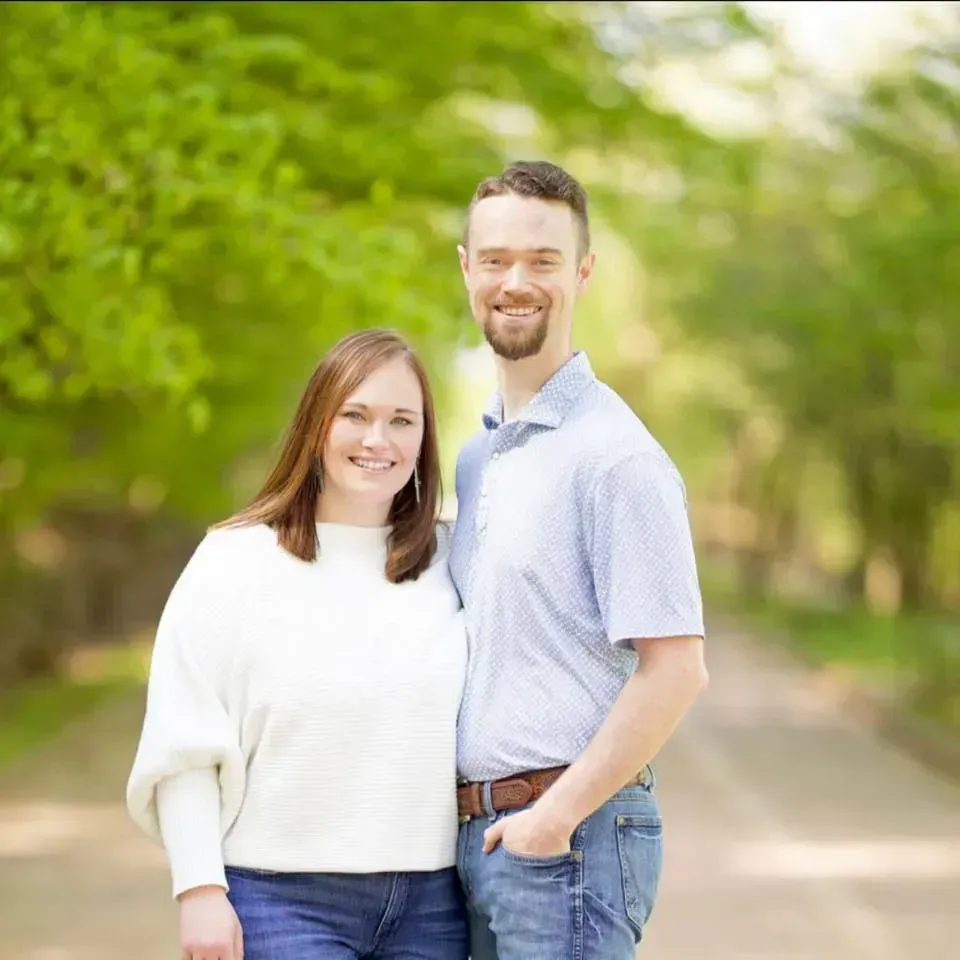 Two people smiling outdoors on a tree-lined path, wearing light-colored casual clothes.
