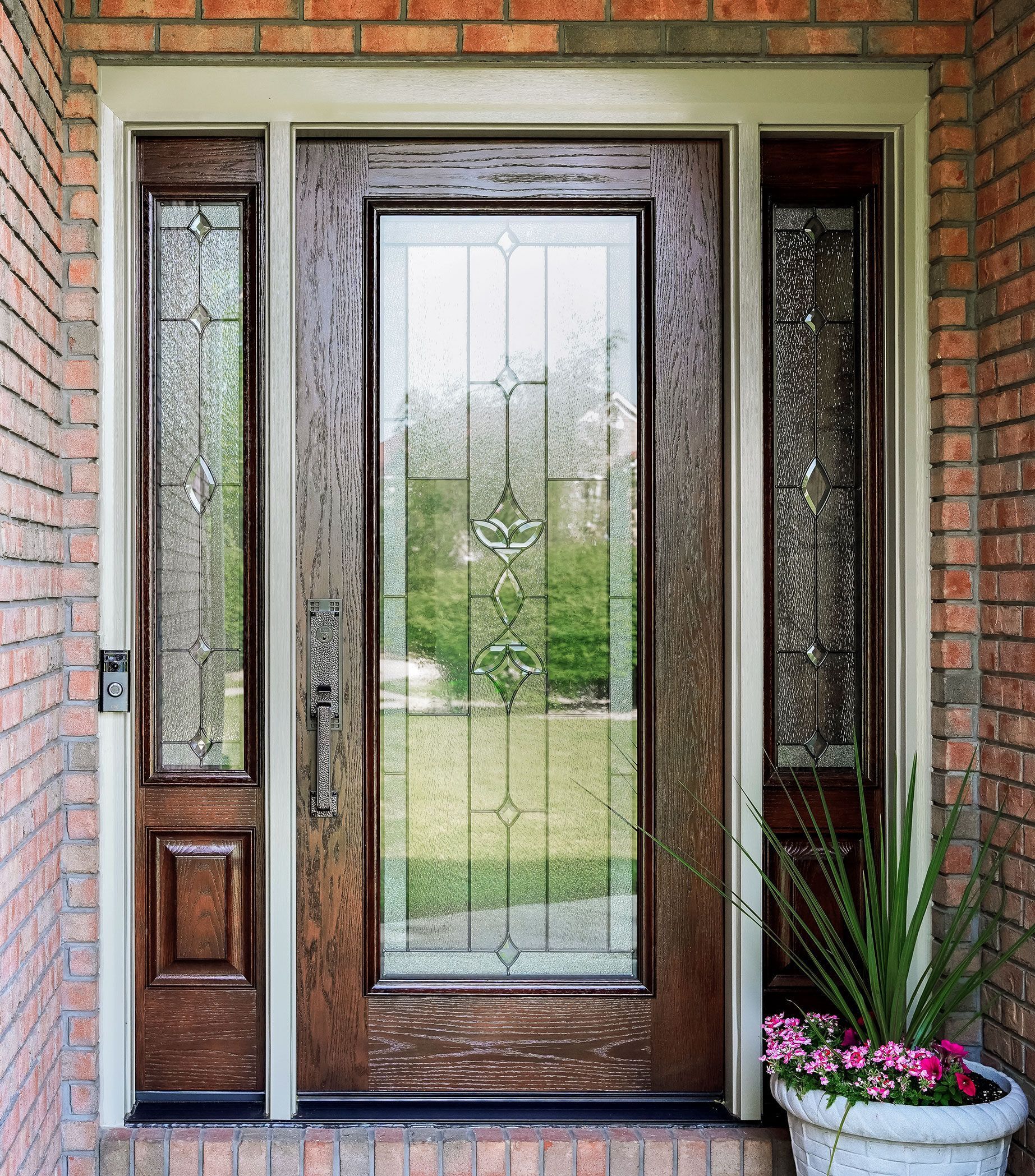 Wooden front door with glass panels and sidelights in a brick entryway; potted plant to the side.