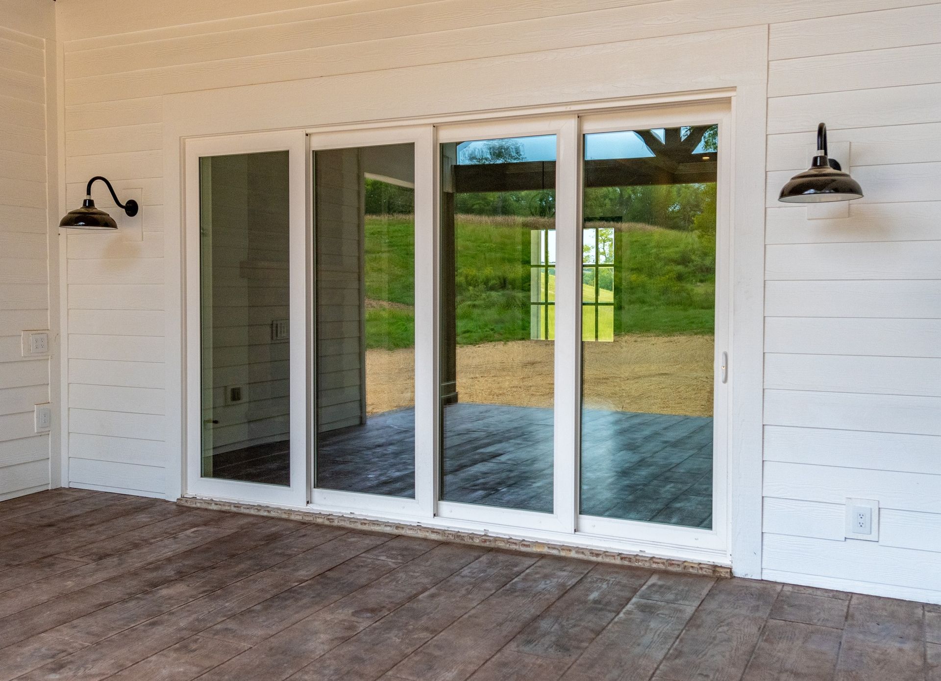 White sliding glass doors on a porch with sconce lighting. Exterior view of a building with a wood deck and a green landscape.