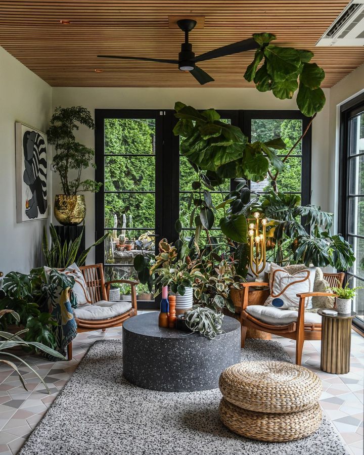 Sunroom with lush plants, wood chairs, black doors, and a gray stone coffee table.