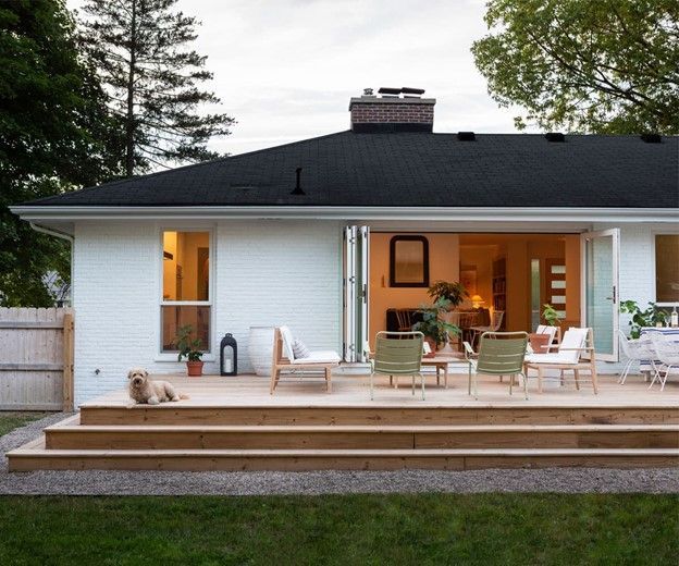 A white house with a wooden deck; bi-fold doors open to reveal interior. Patio furniture and dog on deck.