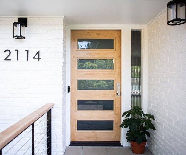Modern wooden front door with glass panels; black address numbers and light fixtures; brick wall.