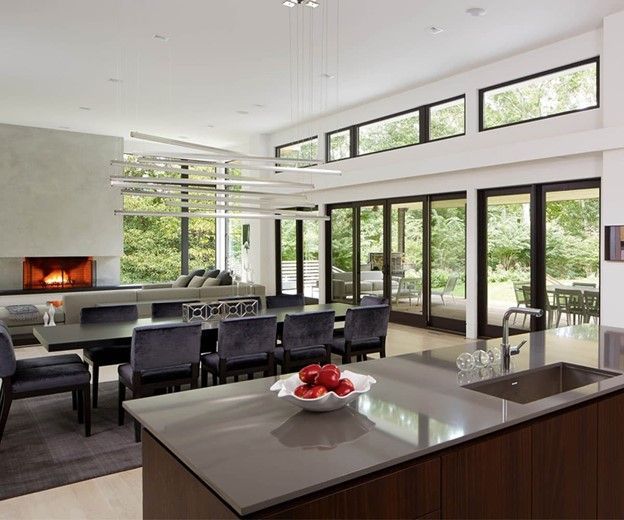 Modern dining and living area with long table, fireplace, and large windows overlooking a green yard.