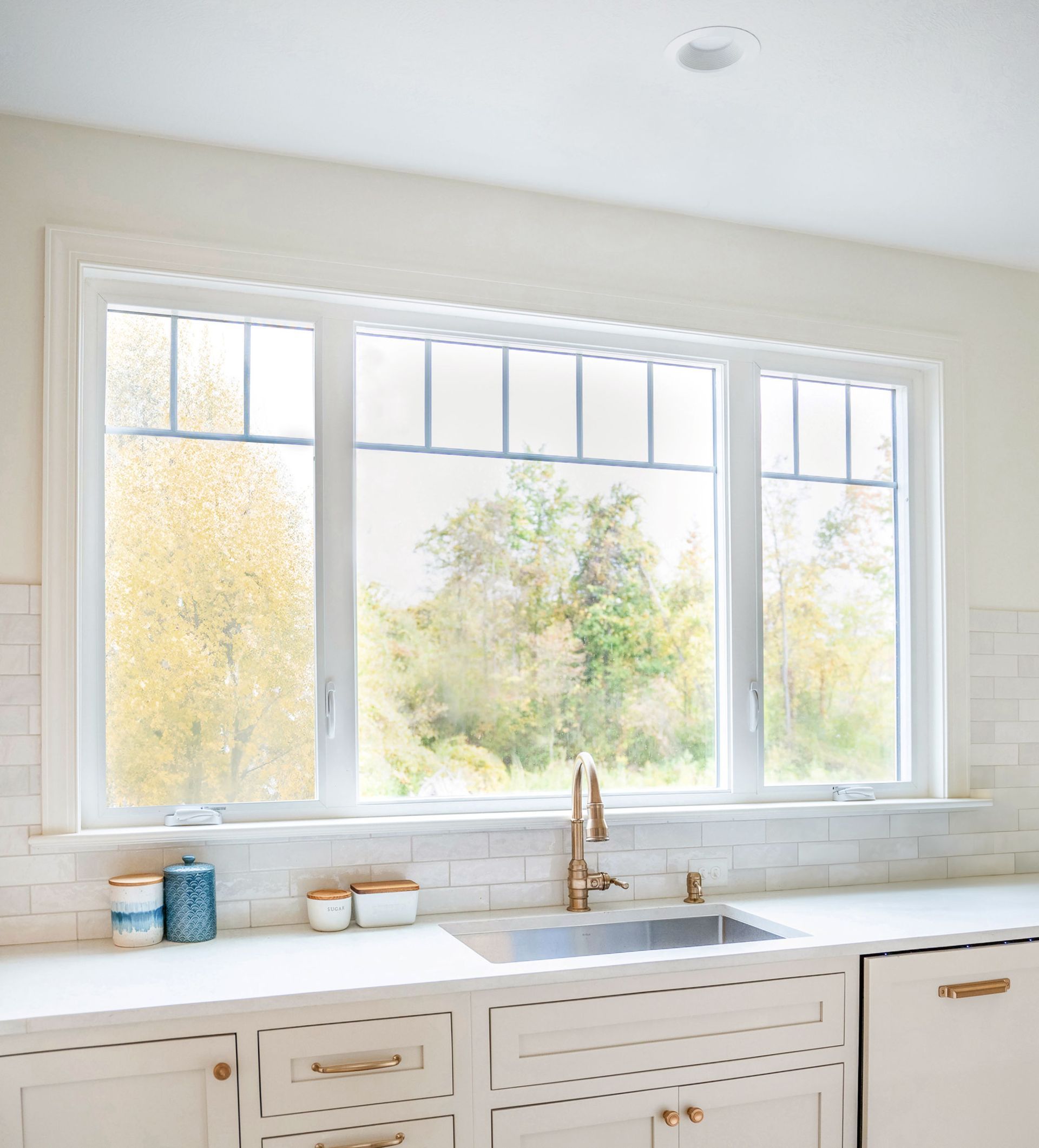 Kitchen window above sink, with white cabinets, gold faucet, and a view of trees.