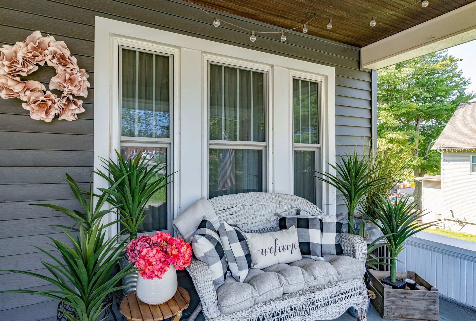 Cozy front porch with white wicker sofa, plants, wreath, and string lights.