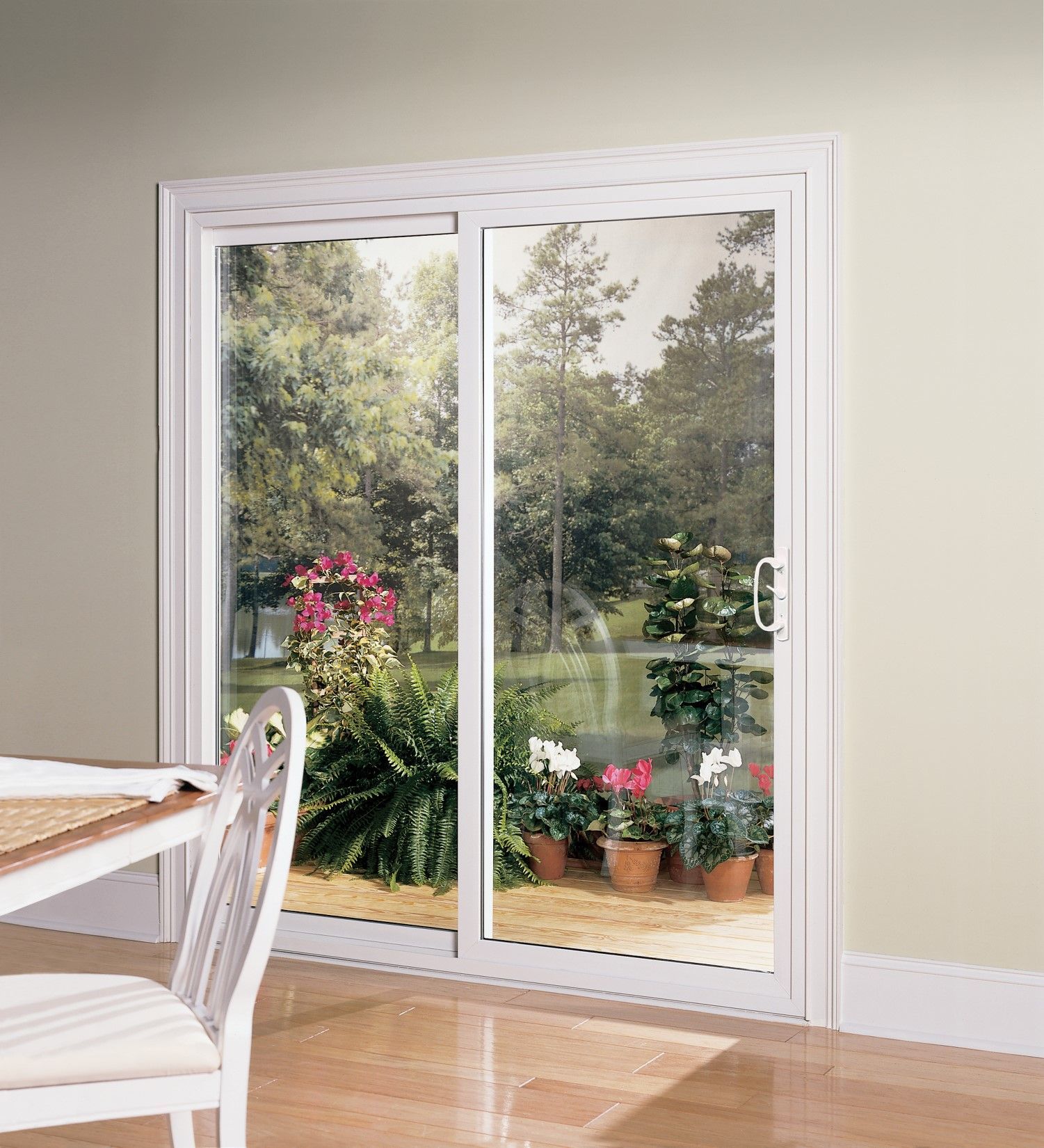 Sliding glass door, white frame, opens to a garden with flowers and trees; a white chair and table are in the foreground.