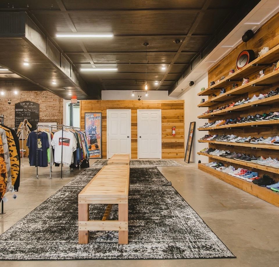 Sneaker store interior with wood shelving displaying shoes, clothing racks, and a bench on a rug.