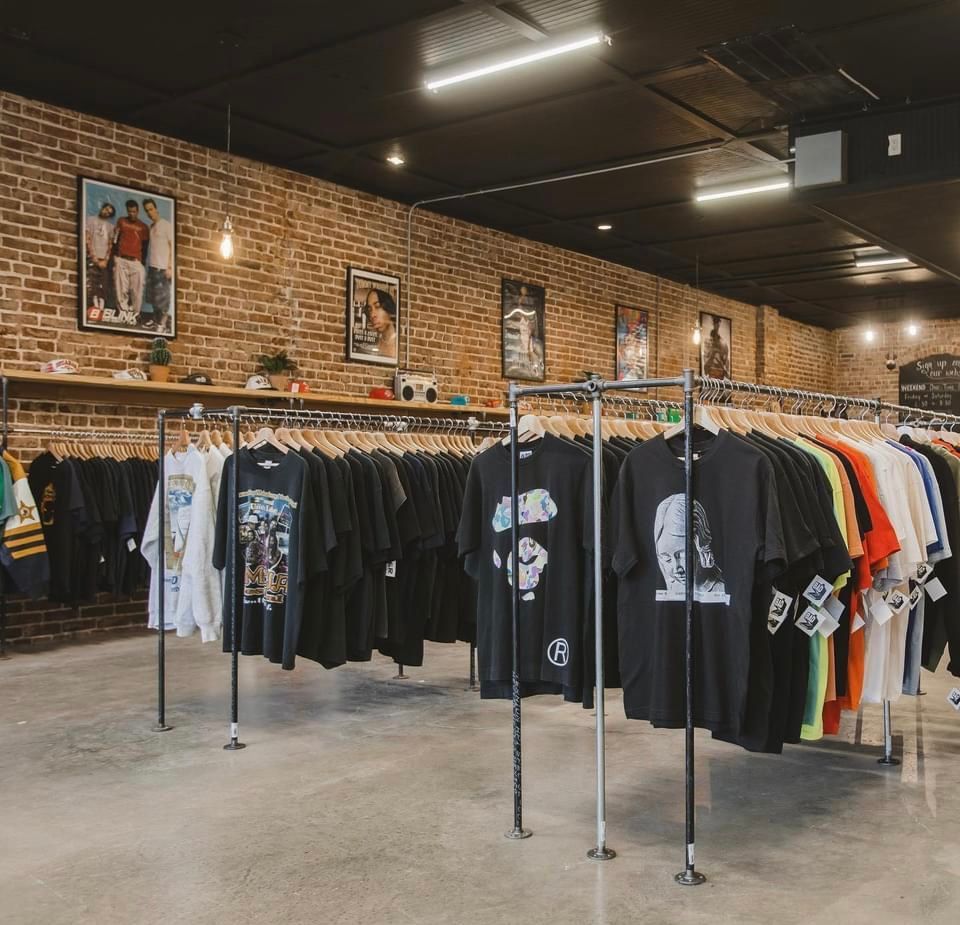 Clothing racks in a brick-walled store, displaying t-shirts, with art on the wall and a dark ceiling.