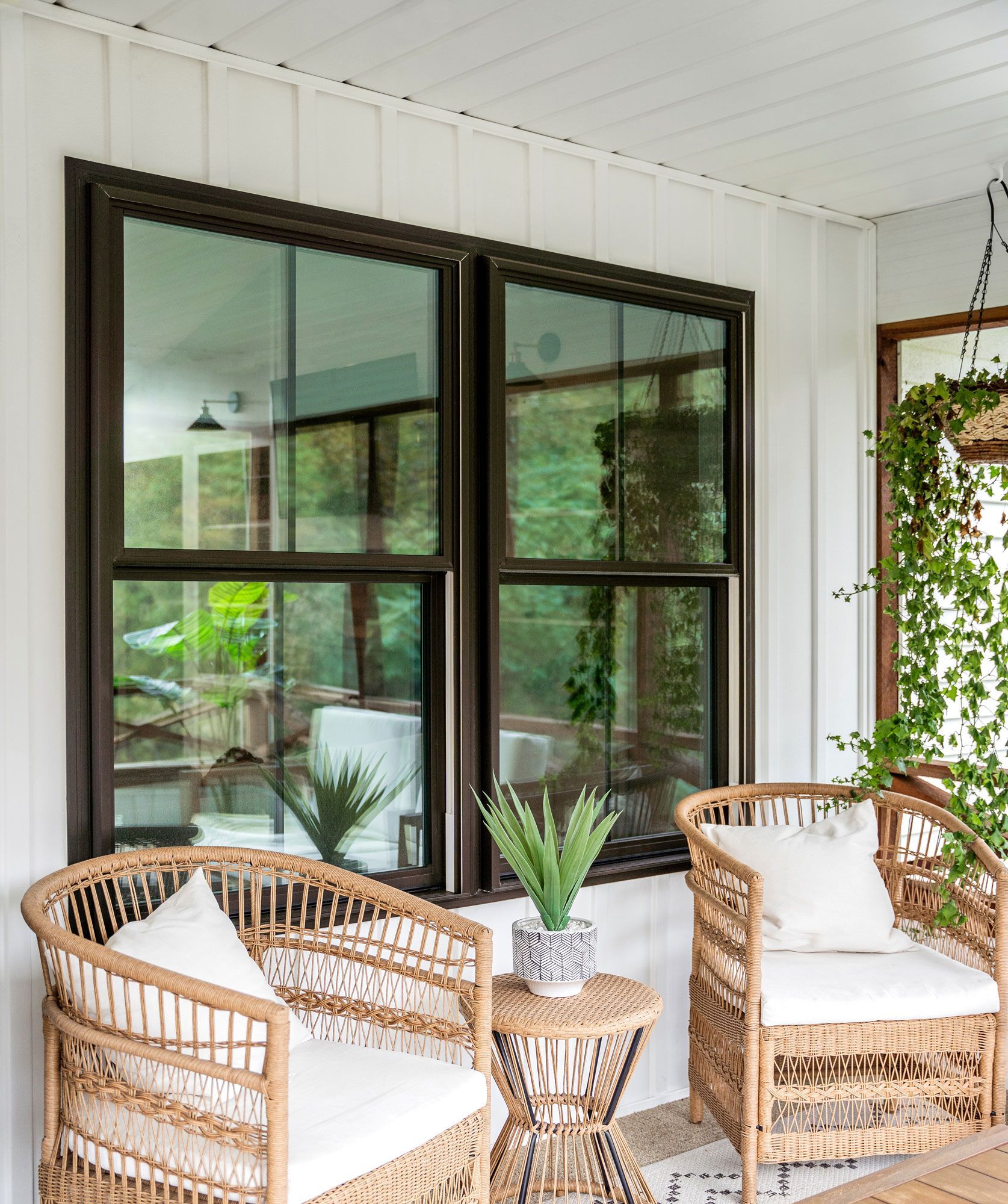 Two wicker chairs on a porch with large windows reflect green foliage, a small table, and a decorative plant.