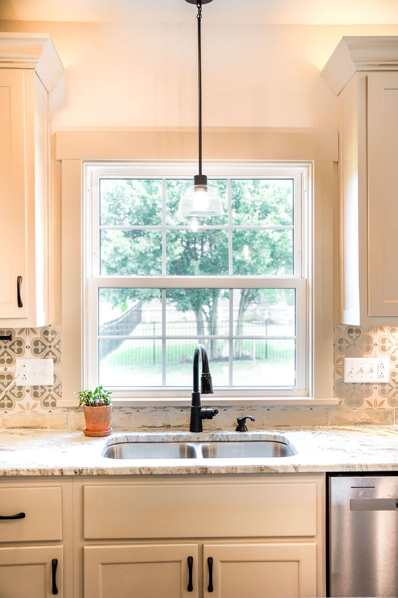 Kitchen with sink, window, light fixture, beige cabinets, granite countertops, and greenery outside the window.