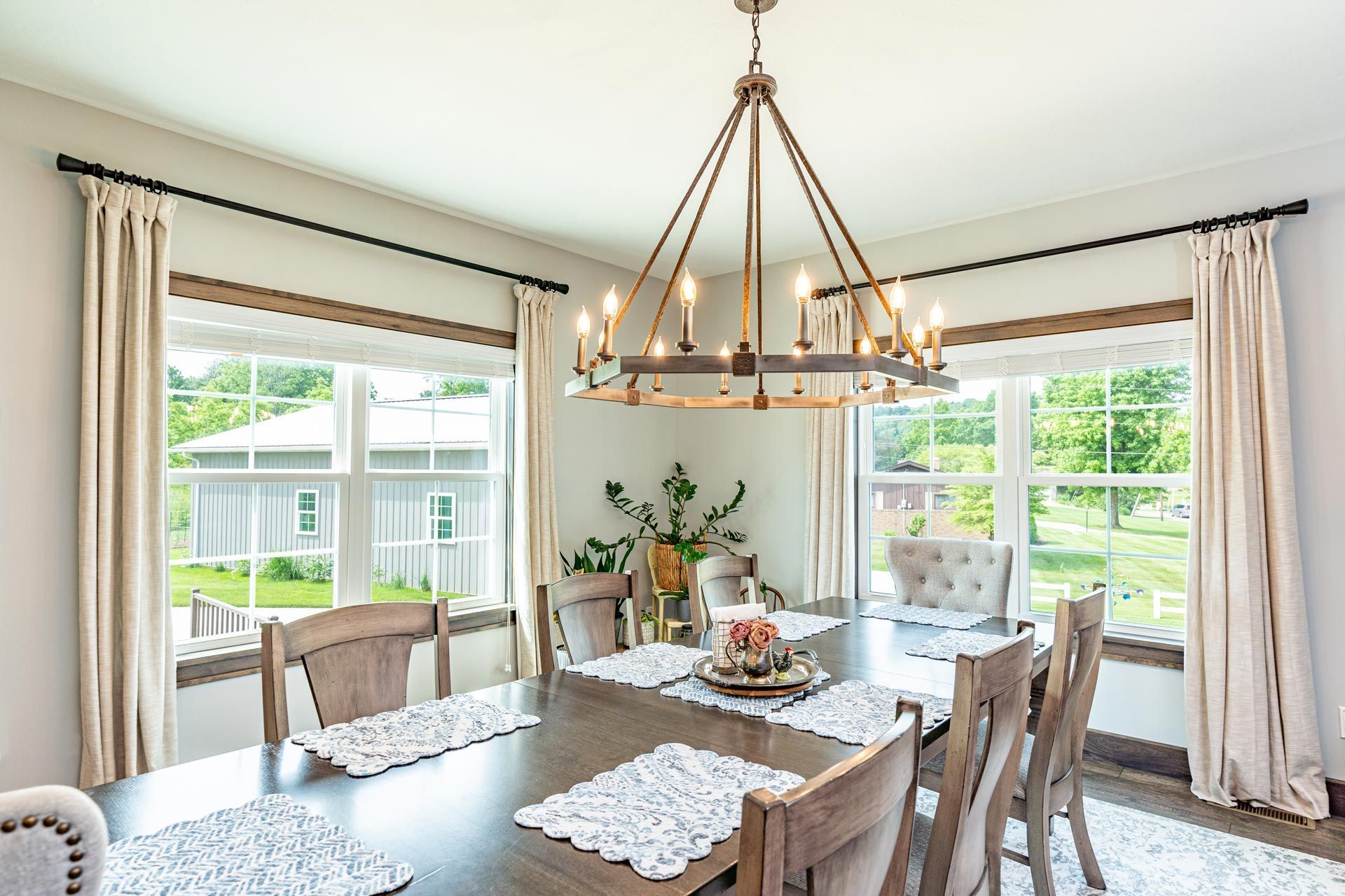 Dining room with a chandelier, wooden table, and chairs near windows with neutral curtains.