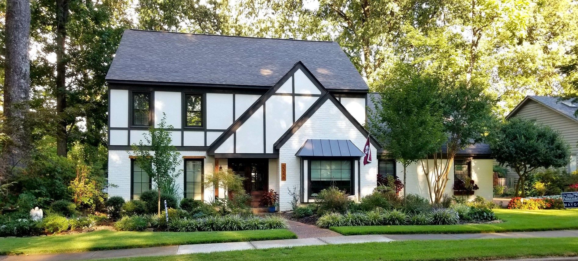 Tudor-style house with white walls, black trim, and a dark roof. It has a well-manicured lawn and flower beds.
