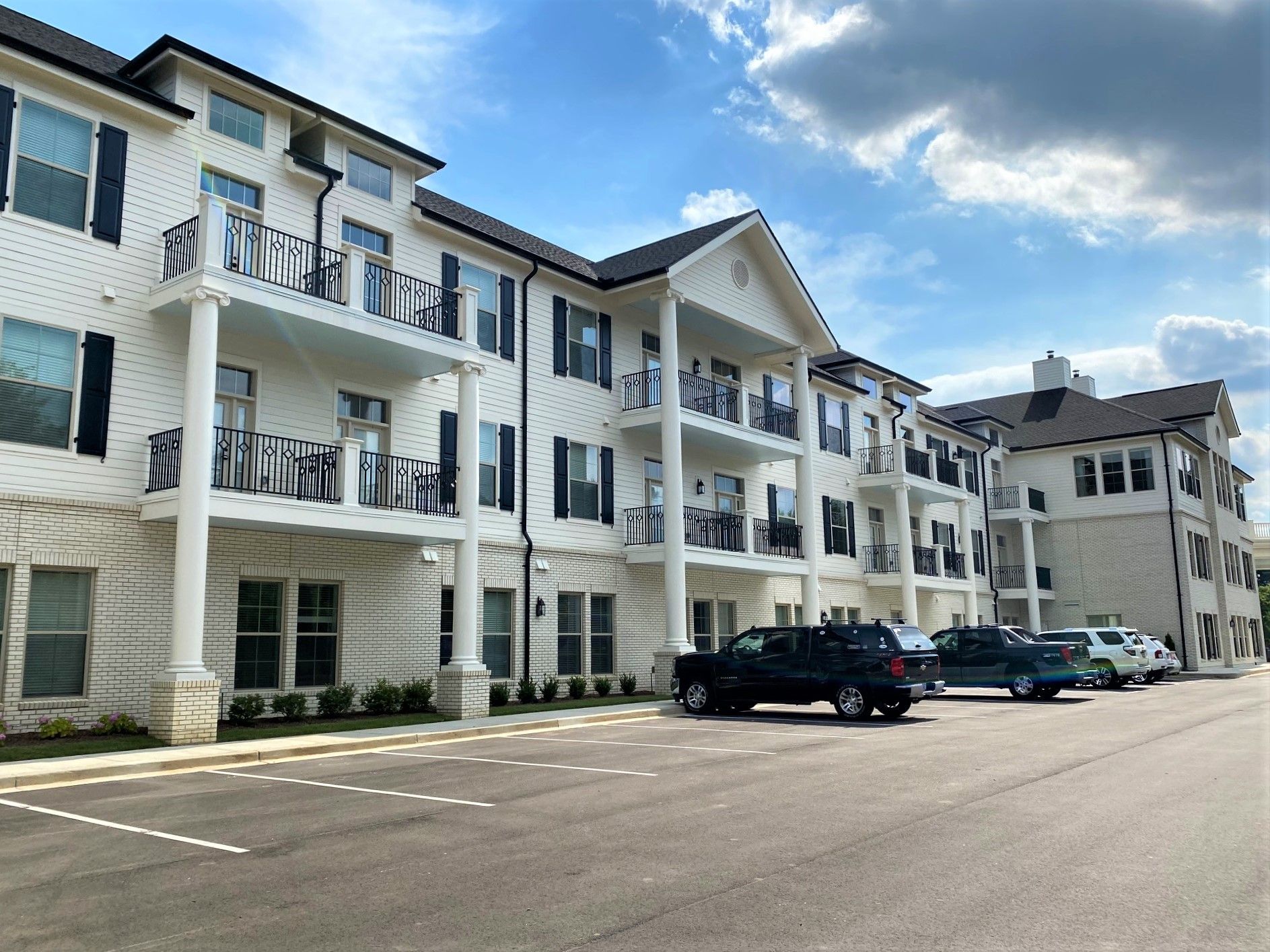 White apartment building with balconies, black shutters, and parked cars on a sunny day.