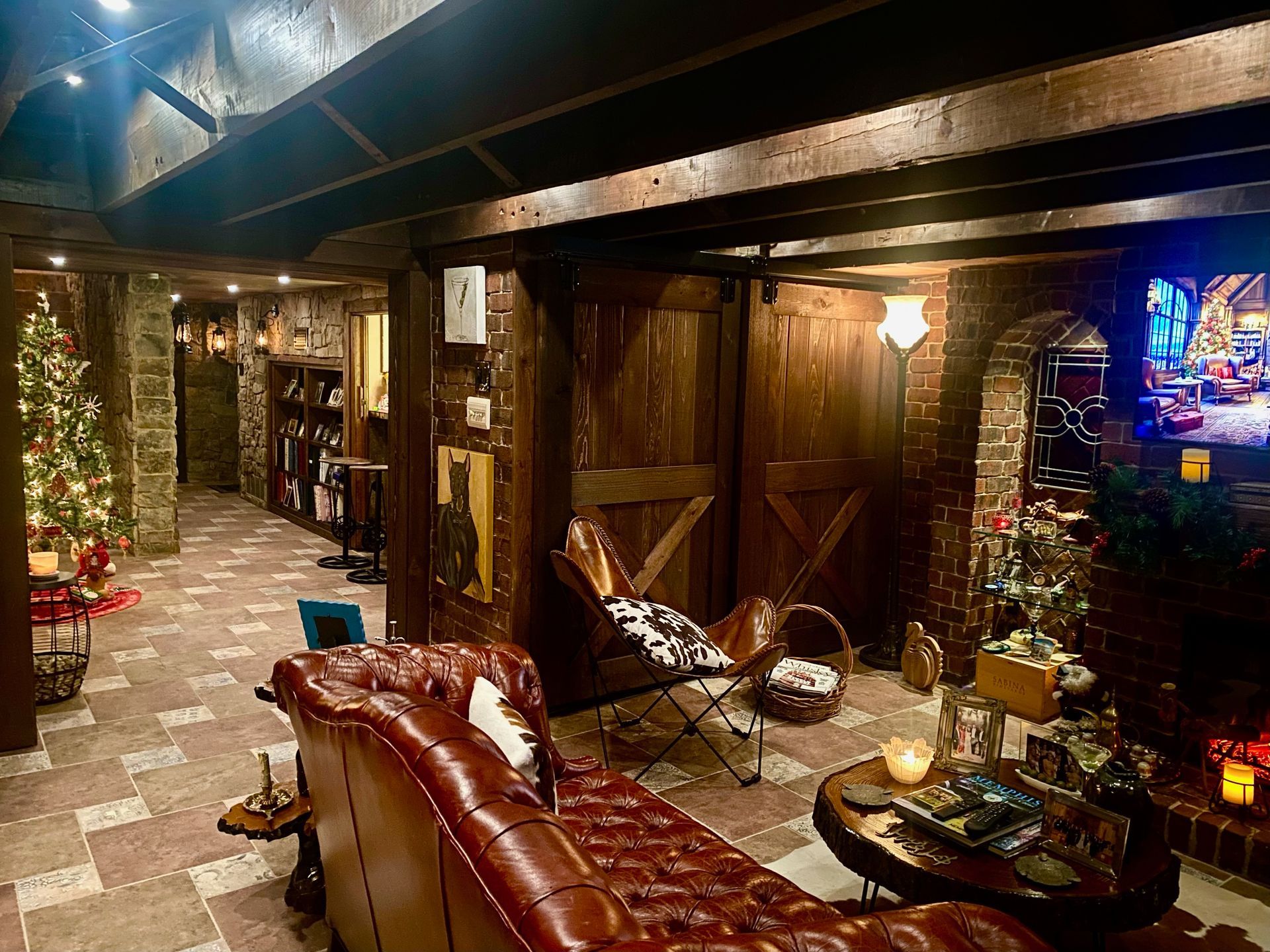 Cozy basement lounge with brick floors, exposed beams, and Christmas decorations; features leather couch and barn doors.