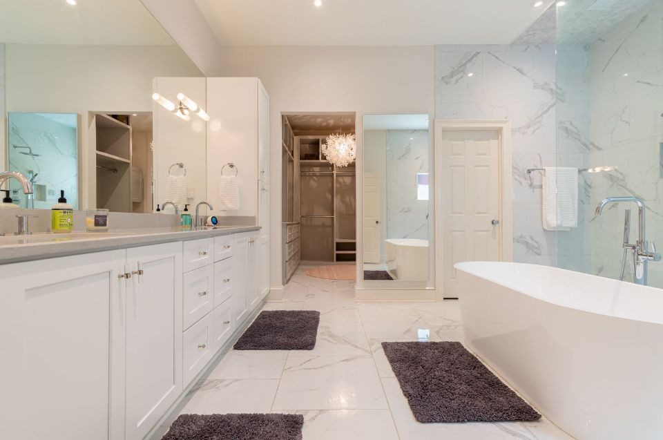 Spacious white bathroom with marble tile, large vanity, freestanding tub, and a walk-in closet in the background.