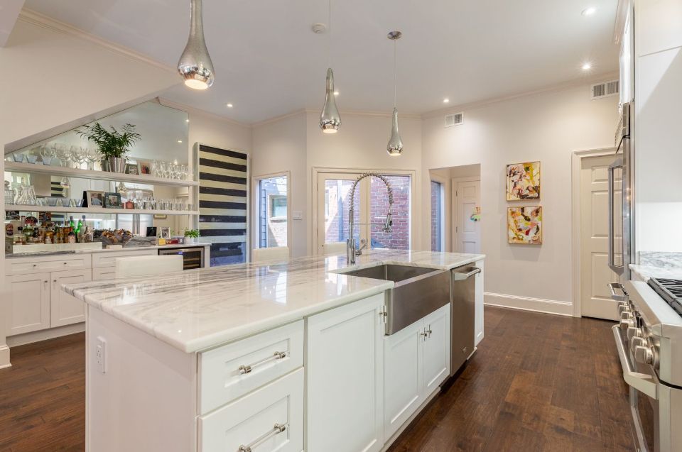 Bright, white kitchen with a large island and stainless steel sink, dark wood floors, and decorative lighting.