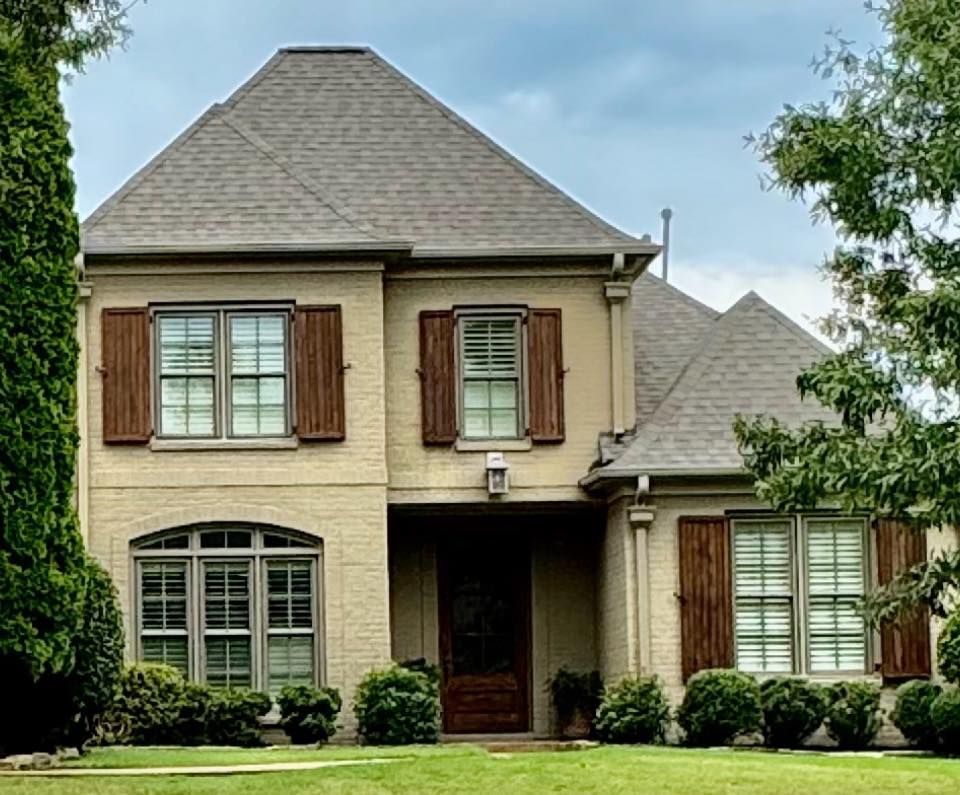 Two-story beige brick house with brown shutters and roof, set on a green lawn with trees.