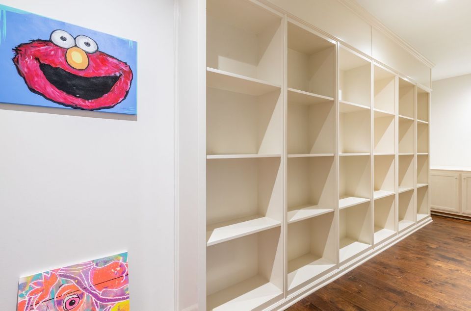 Empty white built-in shelves on a wall, with artwork and wood floor in a brightly lit room.