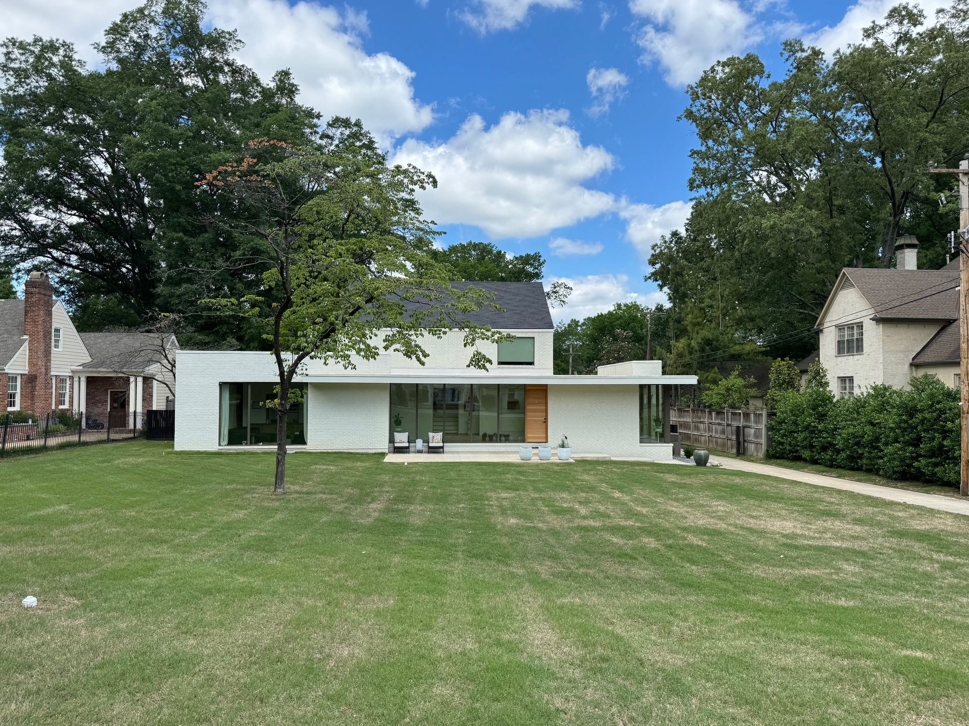 White modern house with flat roof, glass walls, and a small tree in front. Green lawn, blue sky.