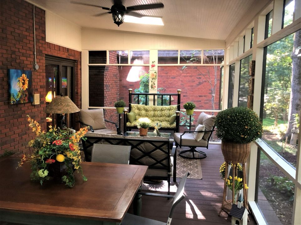 Screened-in porch with brick wall, comfortable seating, dining table, and potted plants. Sunlight streams in through windows.