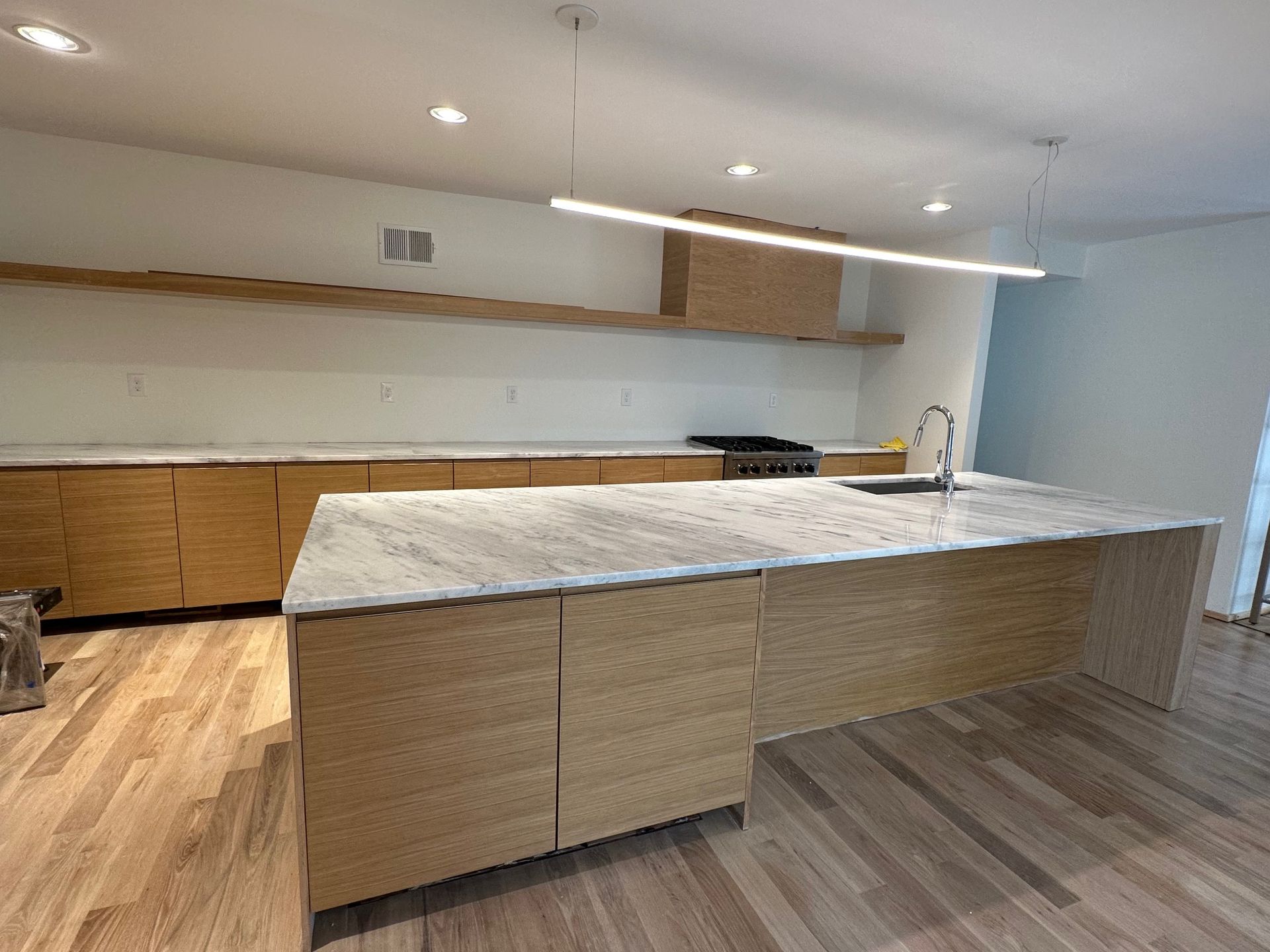Modern kitchen with light wood cabinetry, white countertops, island, and linear pendant light.