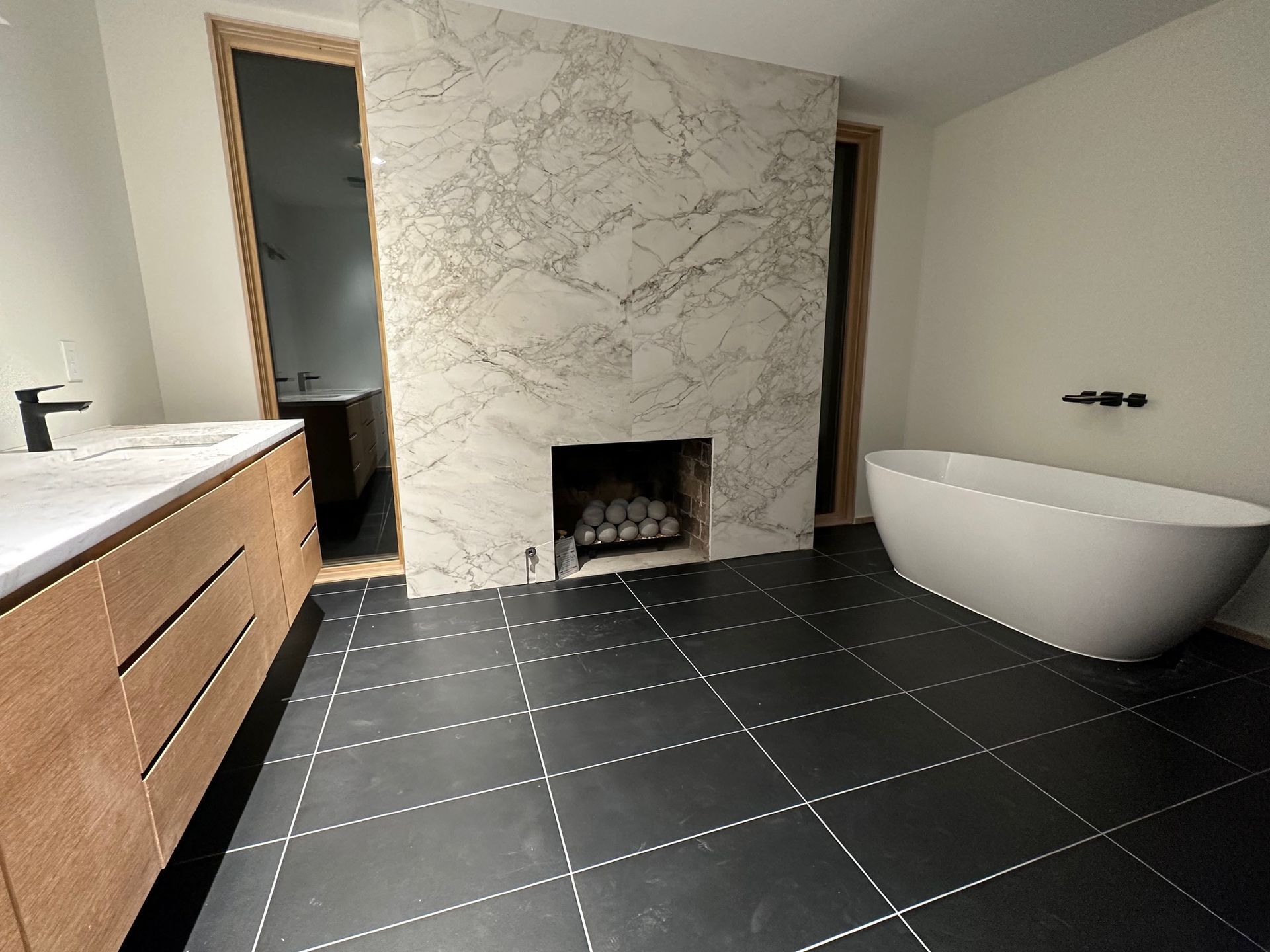 Modern bathroom with a marble fireplace, soaking tub, wood vanity, and black tiled floor.