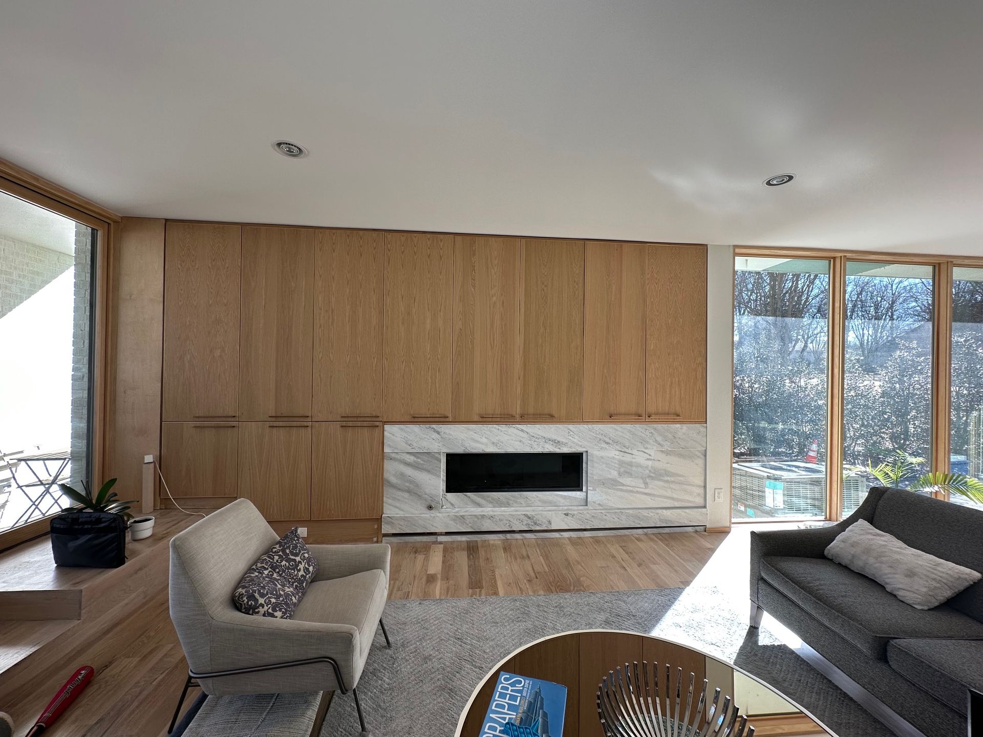 Living room with wood paneling, marble fireplace, and seating; light streams in from large windows.