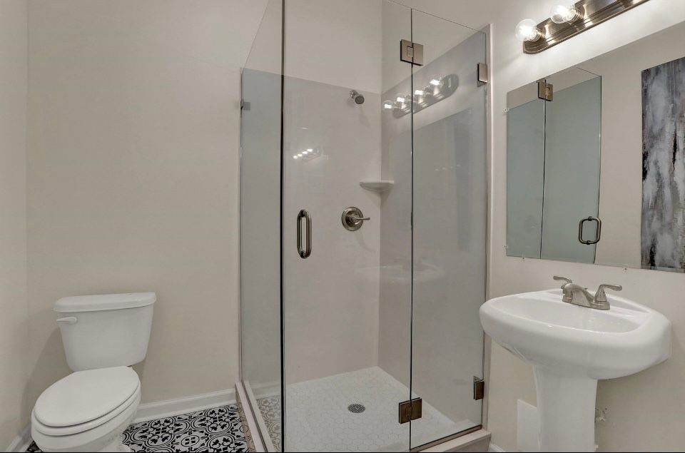Bathroom with a glass shower, pedestal sink, toilet, and vanity mirror. Walls are light-colored.