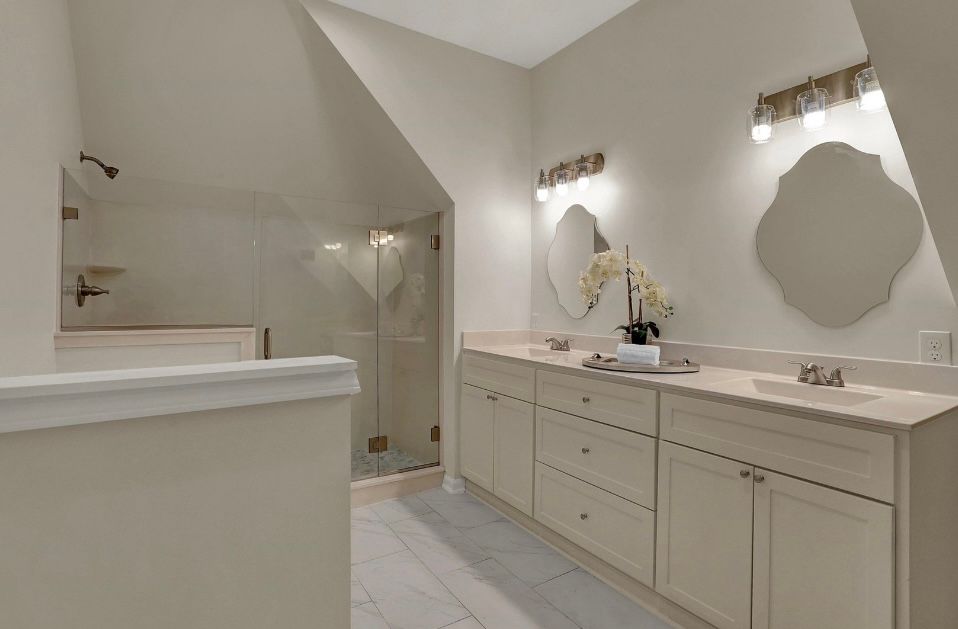Bathroom with beige cabinets, light countertops, and oval mirrors under wall lights. Glass shower.