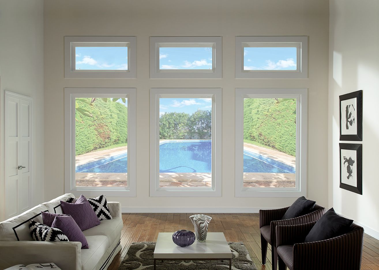 Living room with large windows overlooking a pool. White walls, sofa, chairs, and framed art. Bright sunlight.