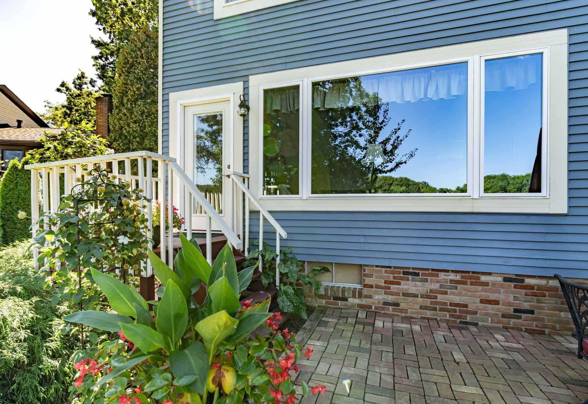 Blue house exterior with white trim, large windows reflecting trees, small patio, and flowering plants.