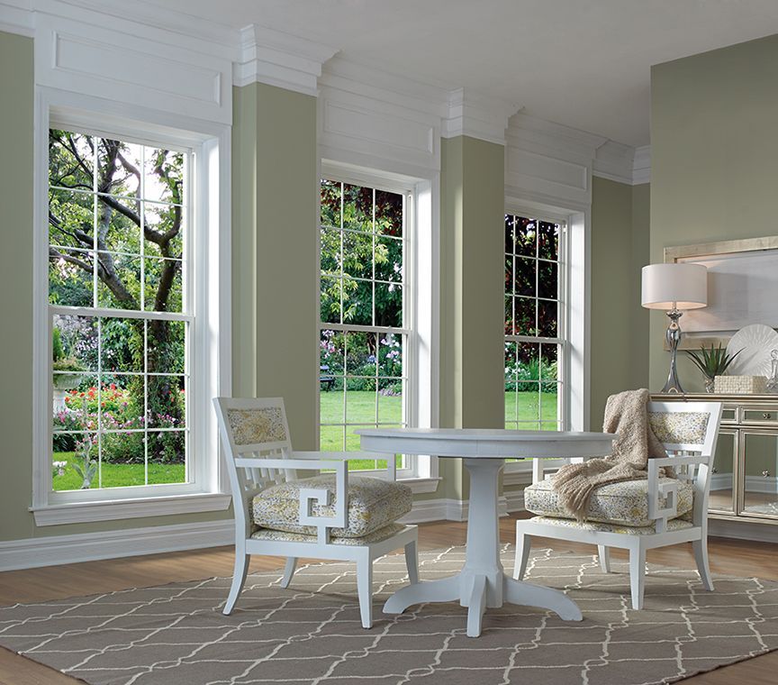 A light-filled room with three large windows overlooking a garden. Table and chairs on a patterned rug.