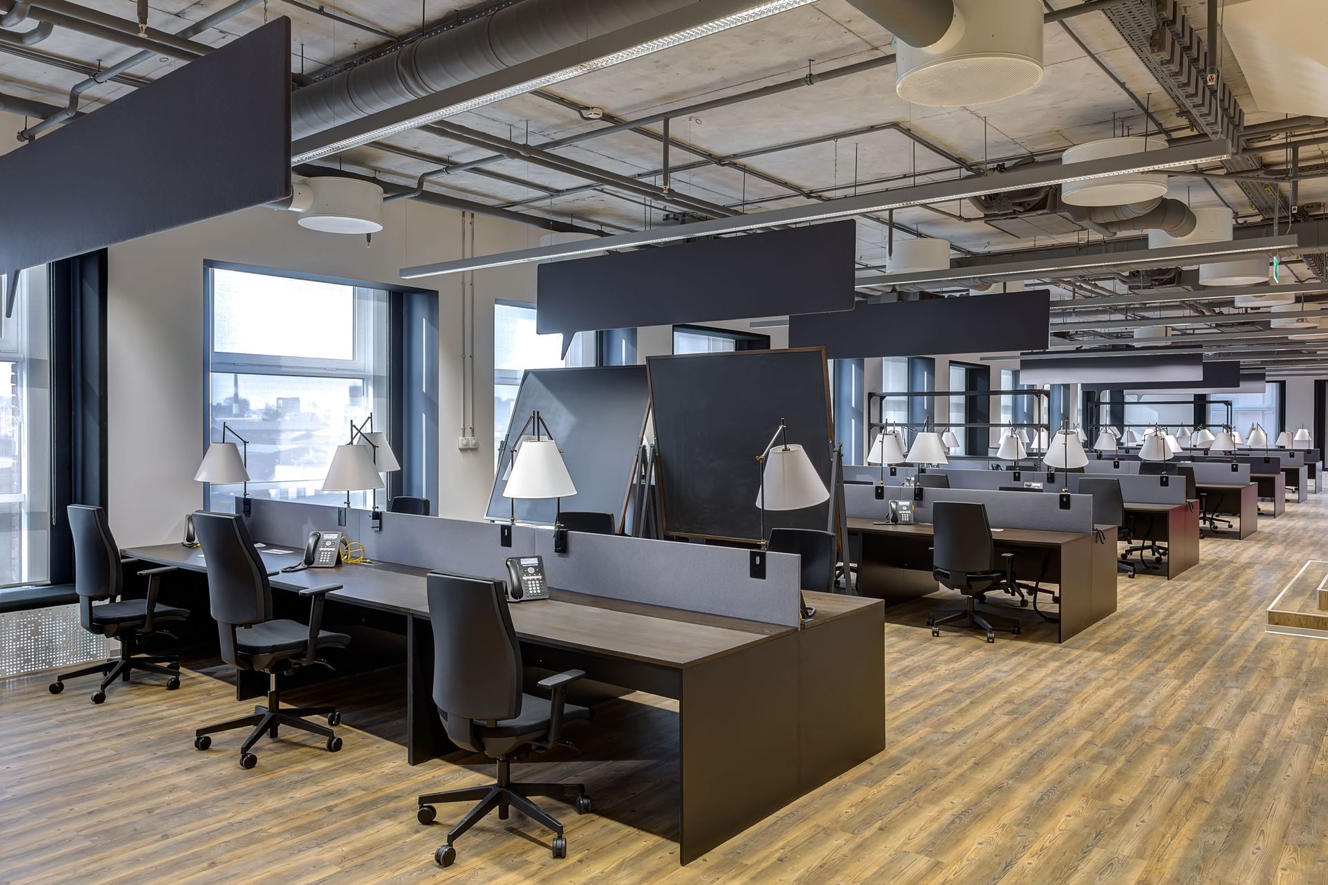 Modern open-plan office with desks, chairs, and lamps. Ceiling has exposed pipes and ductwork.