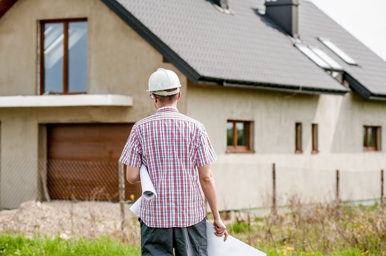 Man in hard hat and plaid shirt, holding blueprints, walks toward a new house.