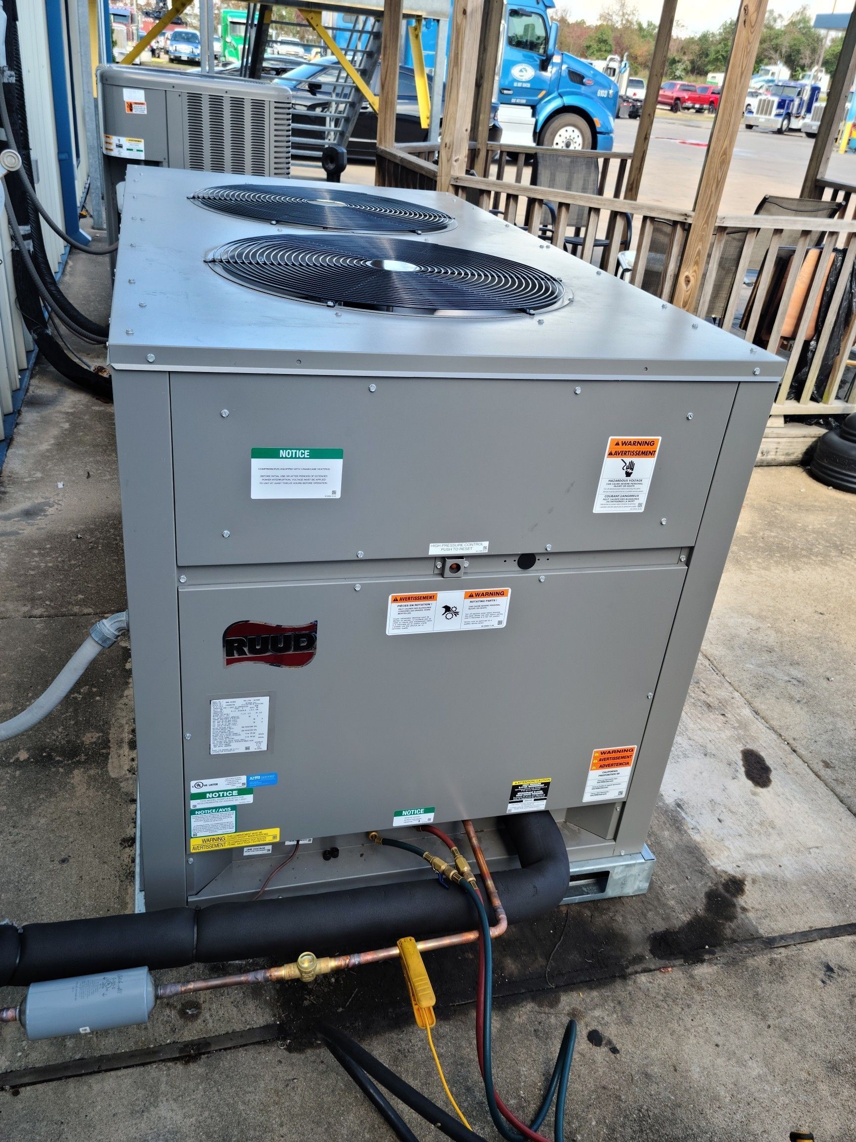 Gray outdoor HVAC unit with fan on top, pipes and cables attached, beside a building walkway