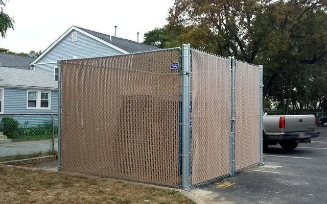 A chain-link privacy fence enclosure with brown slats, situated outdoors in a parking lot next to a residential house.