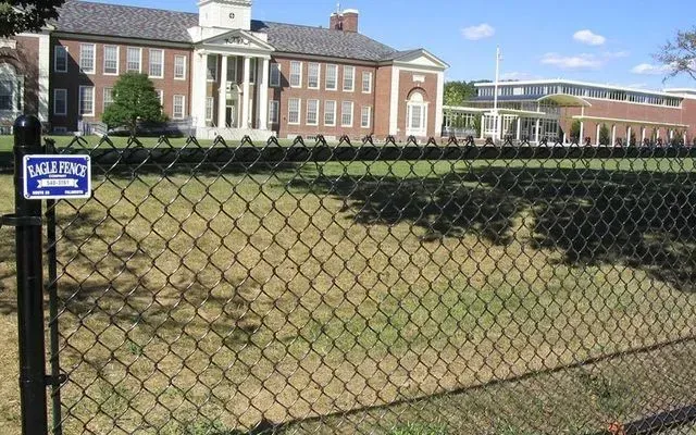 A red brick building with a white columned entrance stands behind a black chain-link fence on a sunny day.