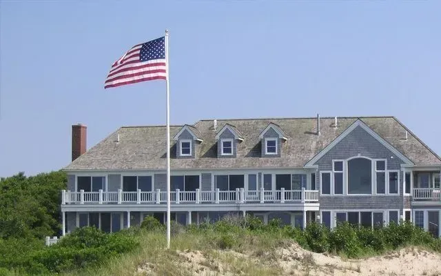 An American flag flies atop a pole in front of a gray, multi-story beach house with shingled siding and dormer windows.