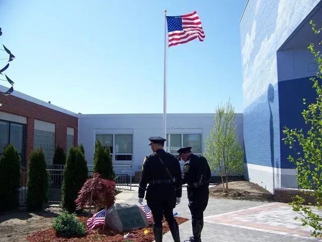 Two officers in uniform stand by a memorial stone and flag at the base of an American flag pole on a sunny day.