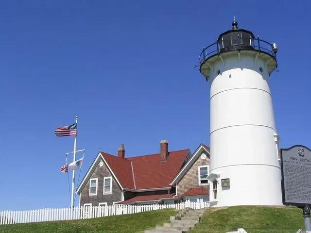 A white lighthouse stands beside a shingled house with a red roof, under a clear blue sky, next to a flag-topped pole.