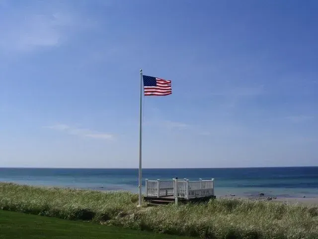 An American flag waves on a flagpole overlooking a calm blue ocean from a grassy dune with a small white deck.